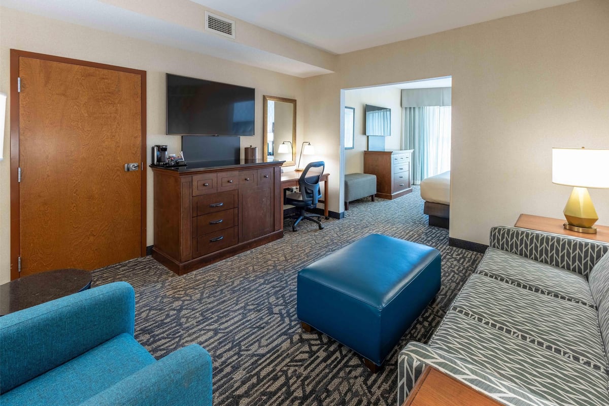 Una habitación de hotel con un escritorio de madera, televisor, sofá, reposapiés y alfombra estampada.