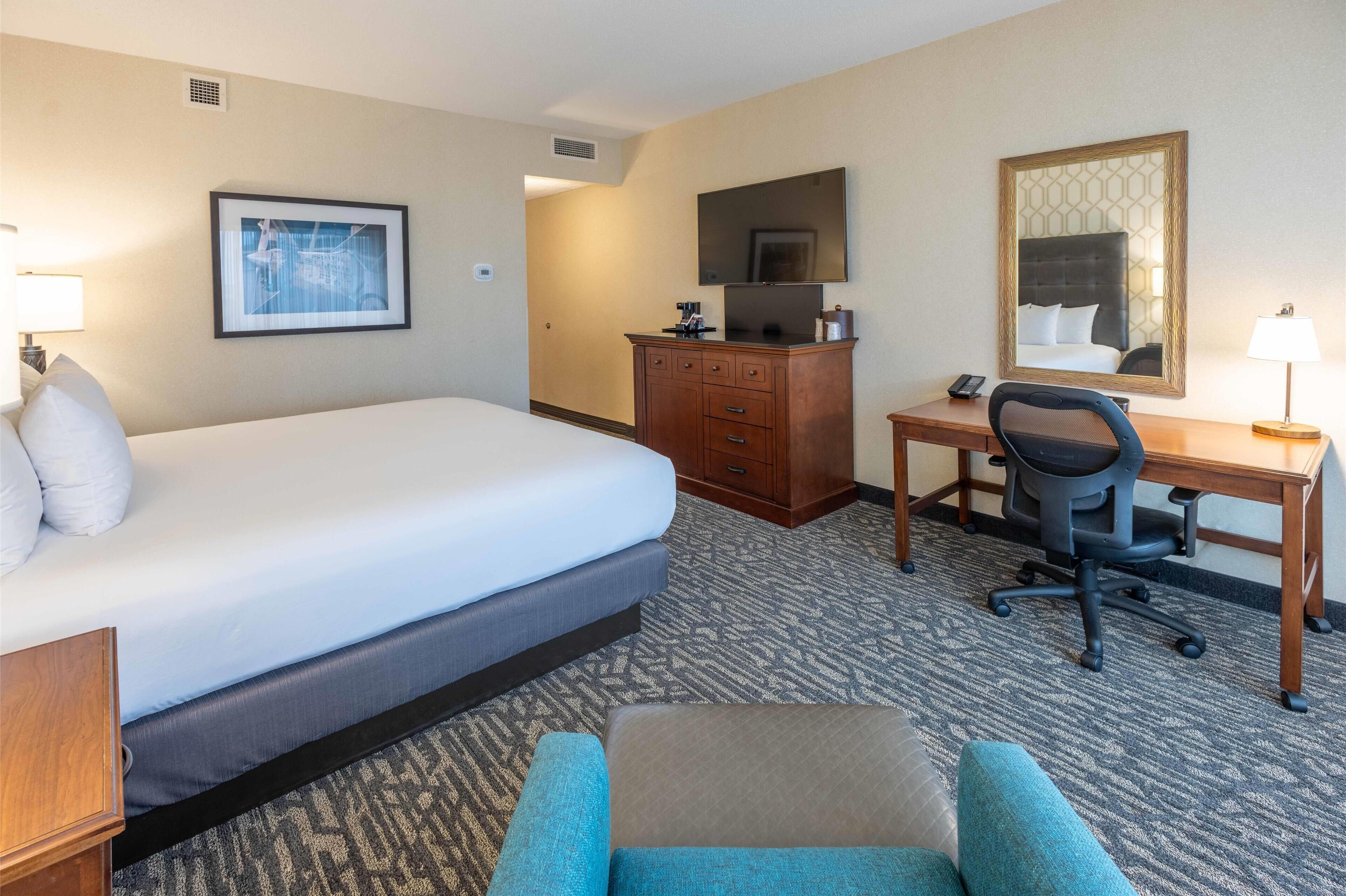 Quarto de hotel com uma cama grande, uma escrivaninha com cadeira, cómoda, espelho e um quadro na parede.