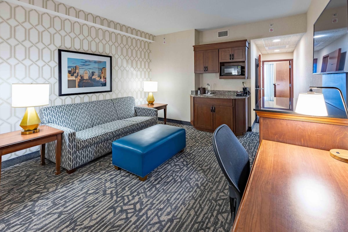 Habitación de hotel con un sofá estampado, un taburete azul, un escritorio de madera, una kitchenette y un cuadro con una vista urbana enmarcada en la pared.