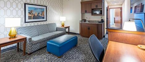 A hotel room with a patterned sofa, a blue ottoman, a wooden desk, a kitchenette, and a framed cityscape picture on the wall.