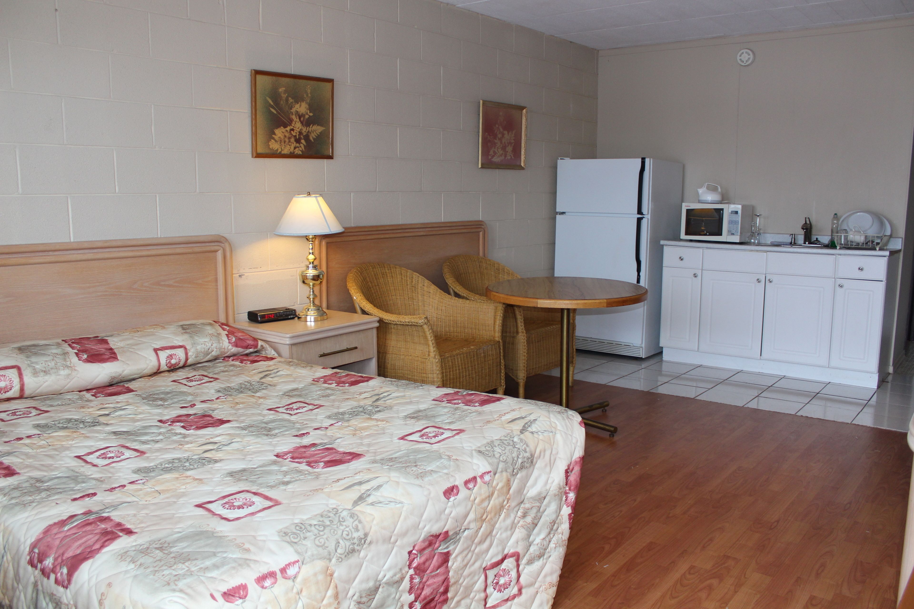 A bedroom with a bed, a nightstand, a lamp, a chair, a small table, a microwave, and a refrigerator.