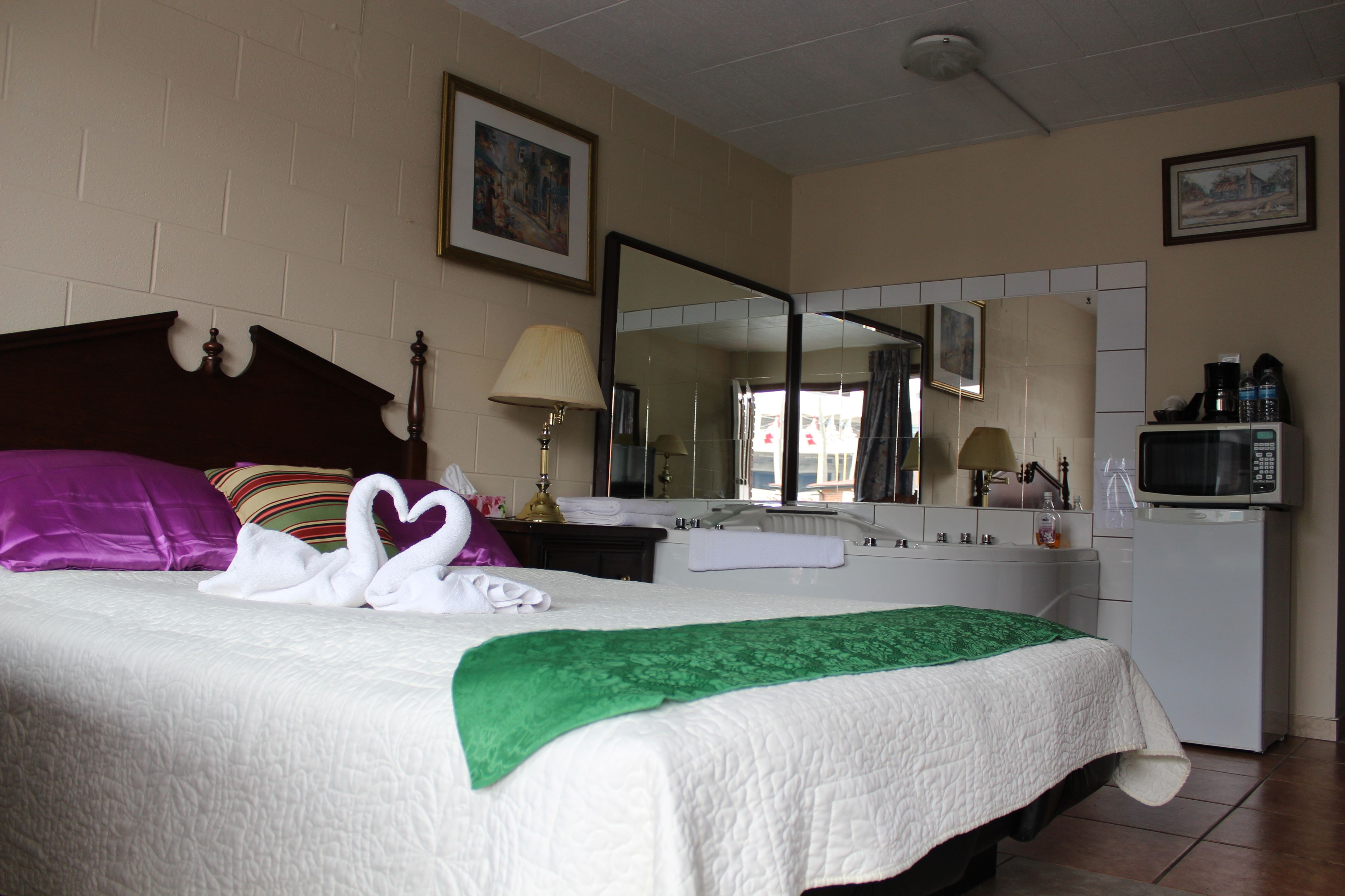 Habitación de hotel con cama, una manta verde, colcha blanca acolchada, almohada morada, almohada a rayas, toalla blanca, lámpara, espejo, microondas y lavamanos.