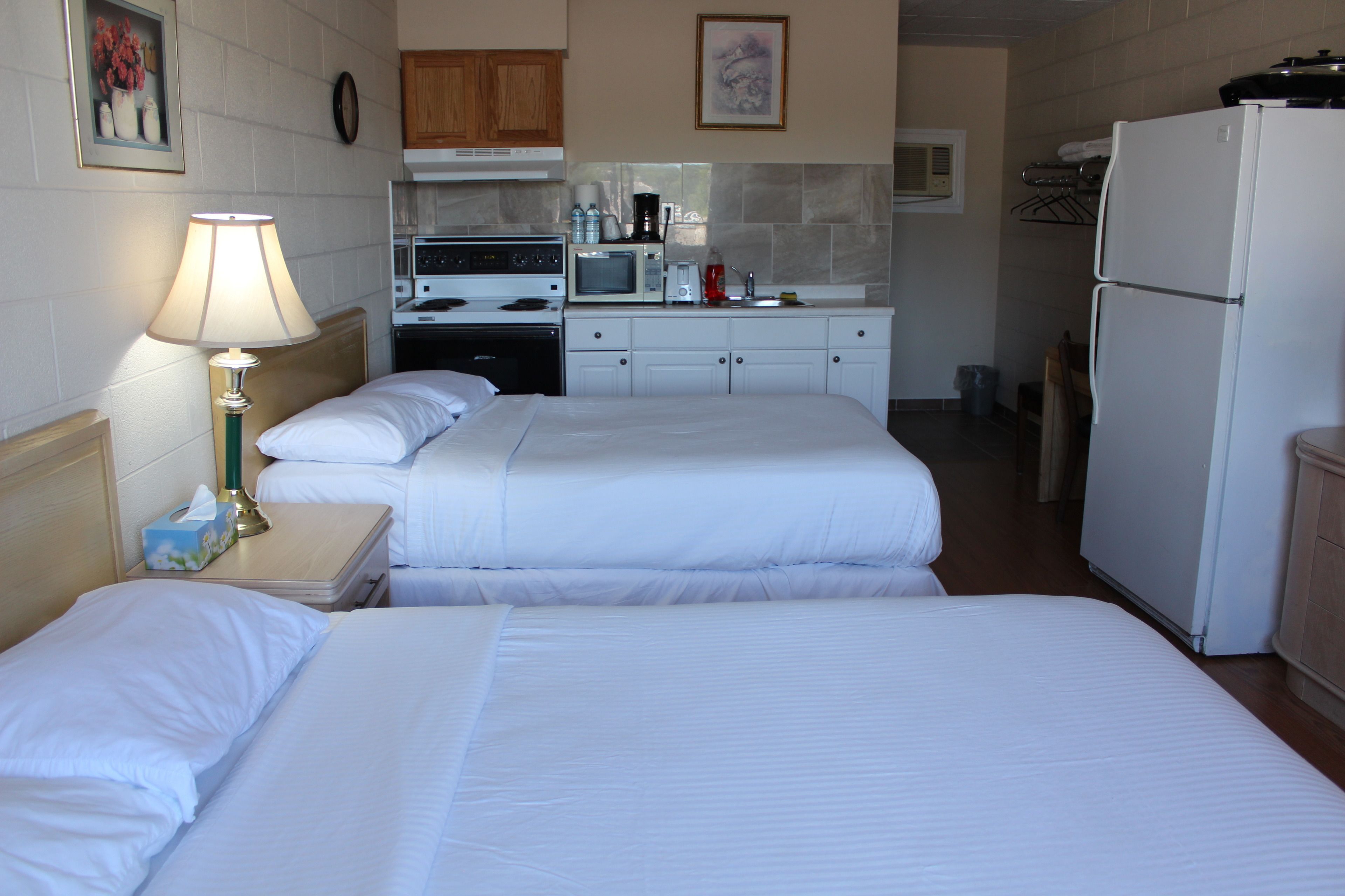 A hotel room with two beds, a nightstand with a lamp, a kitchenette with a stove, microwave, and refrigerator.