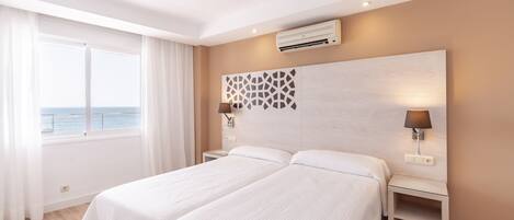 Hotelkamer met twee bedden, een groot raam met gordijnen, een luchtverkoeler aan de muur en twee bedlampjes.