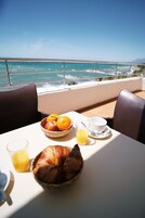 Una mesa con alimentos para el desayuno, un vaso de jugo de naranja y un croissant en un balcón con vista al océano.