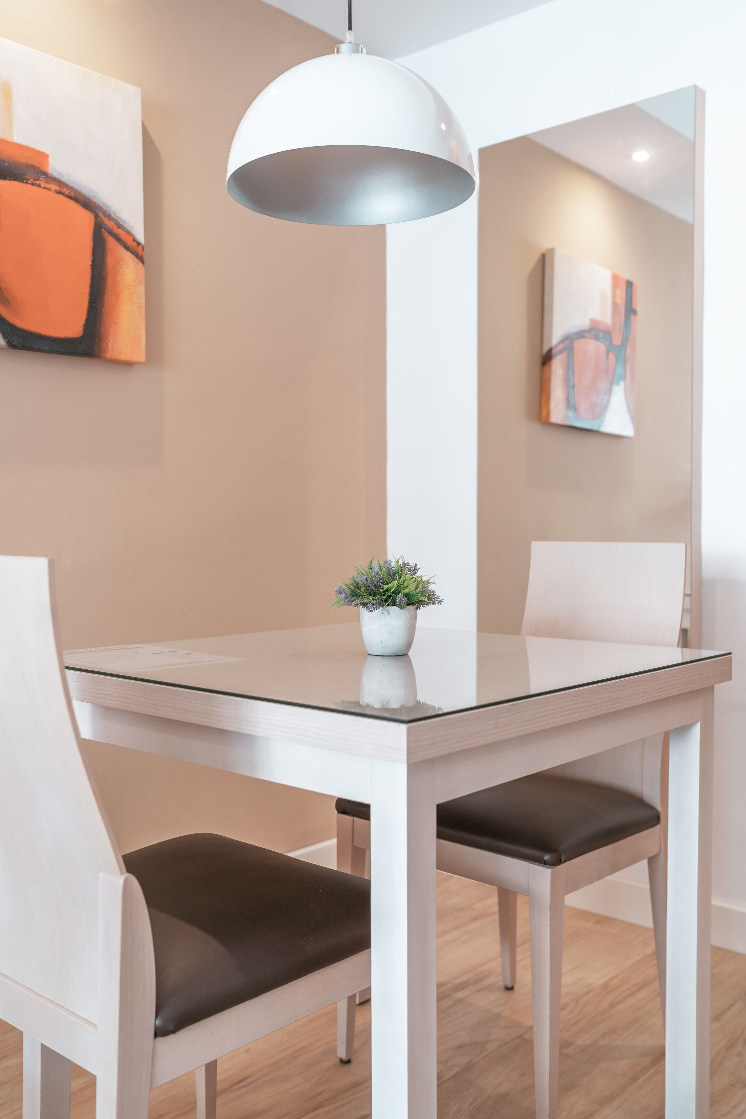 A modern dining area with a glass table, white chairs, a potted plant, and abstract art on the walls.