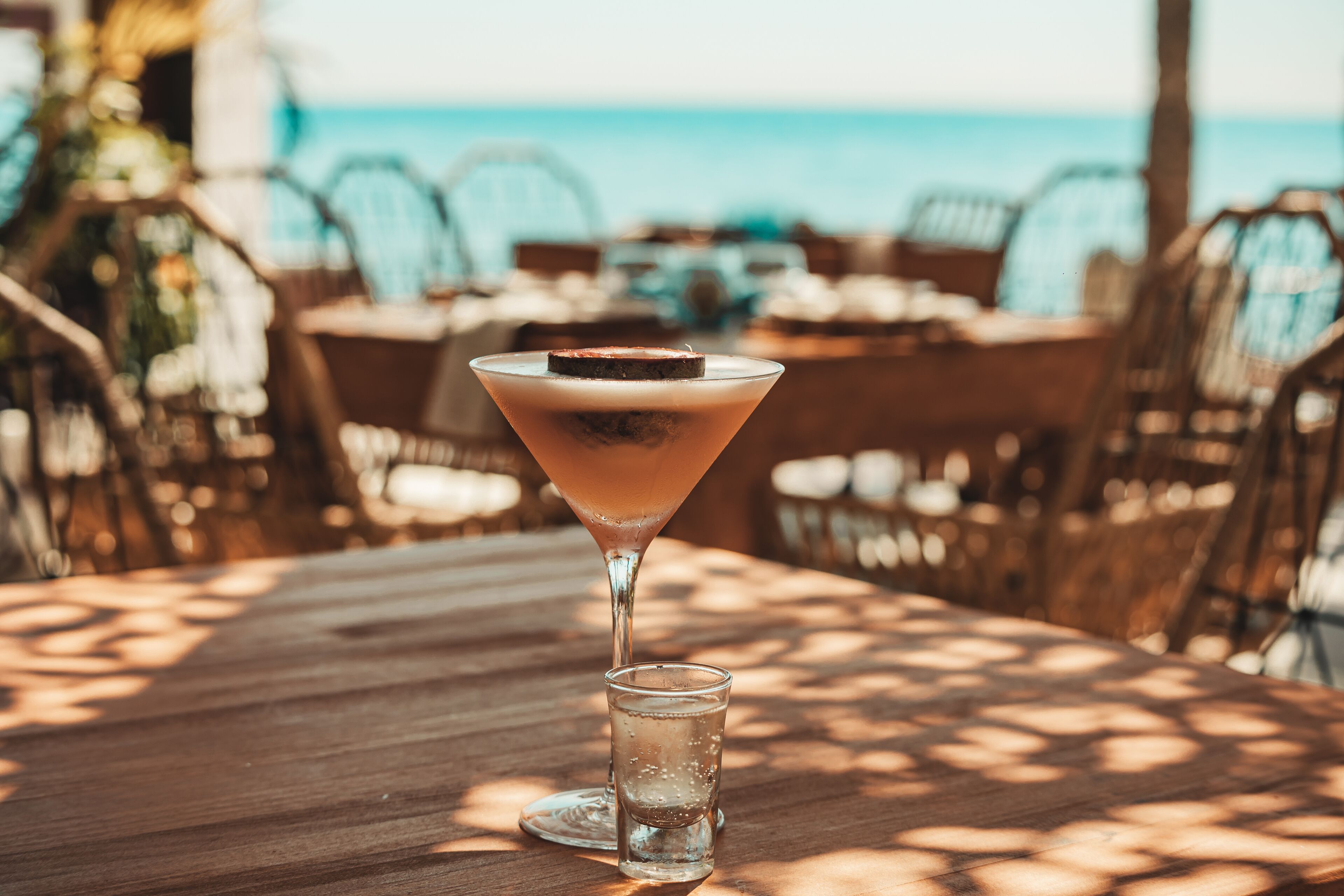 Una copa de martini con una guarnición oscura sobre una mesa de madera, un vaso para chupitos a su lado, y una vista al océano de fondo.