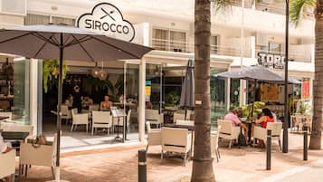 Ett utomhusområde med ett kafé, bord och stolar, parasoller och en skylt med texten ”Sirocco”.