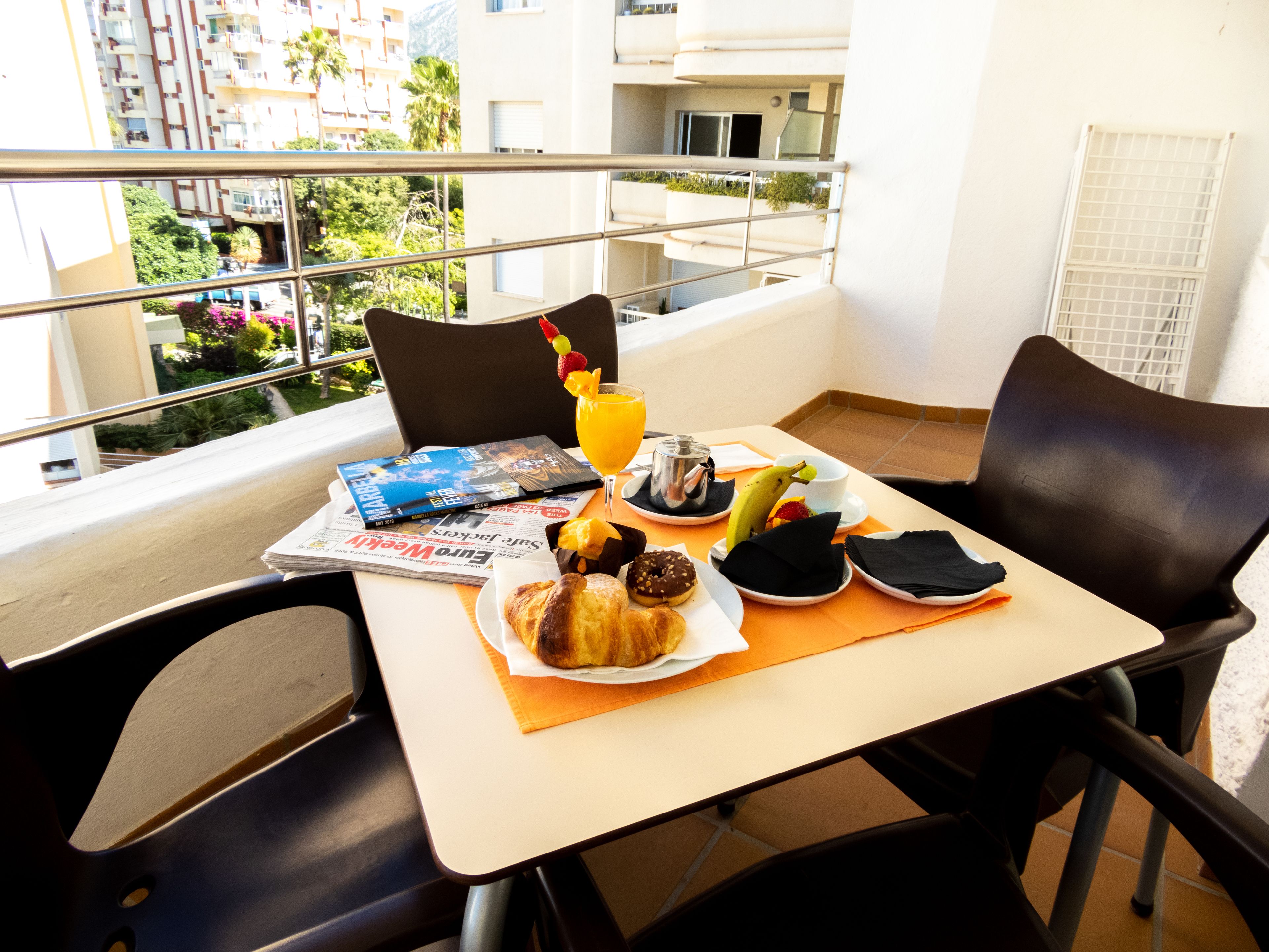 Un tavolo da balcone con articoli per la colazione, una rivista e vista su edifici e spazi verdi.
