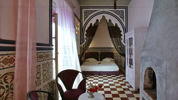 A room with a checkered floor, a bed with a canopy, a chair, and a table with a vase of flowers.