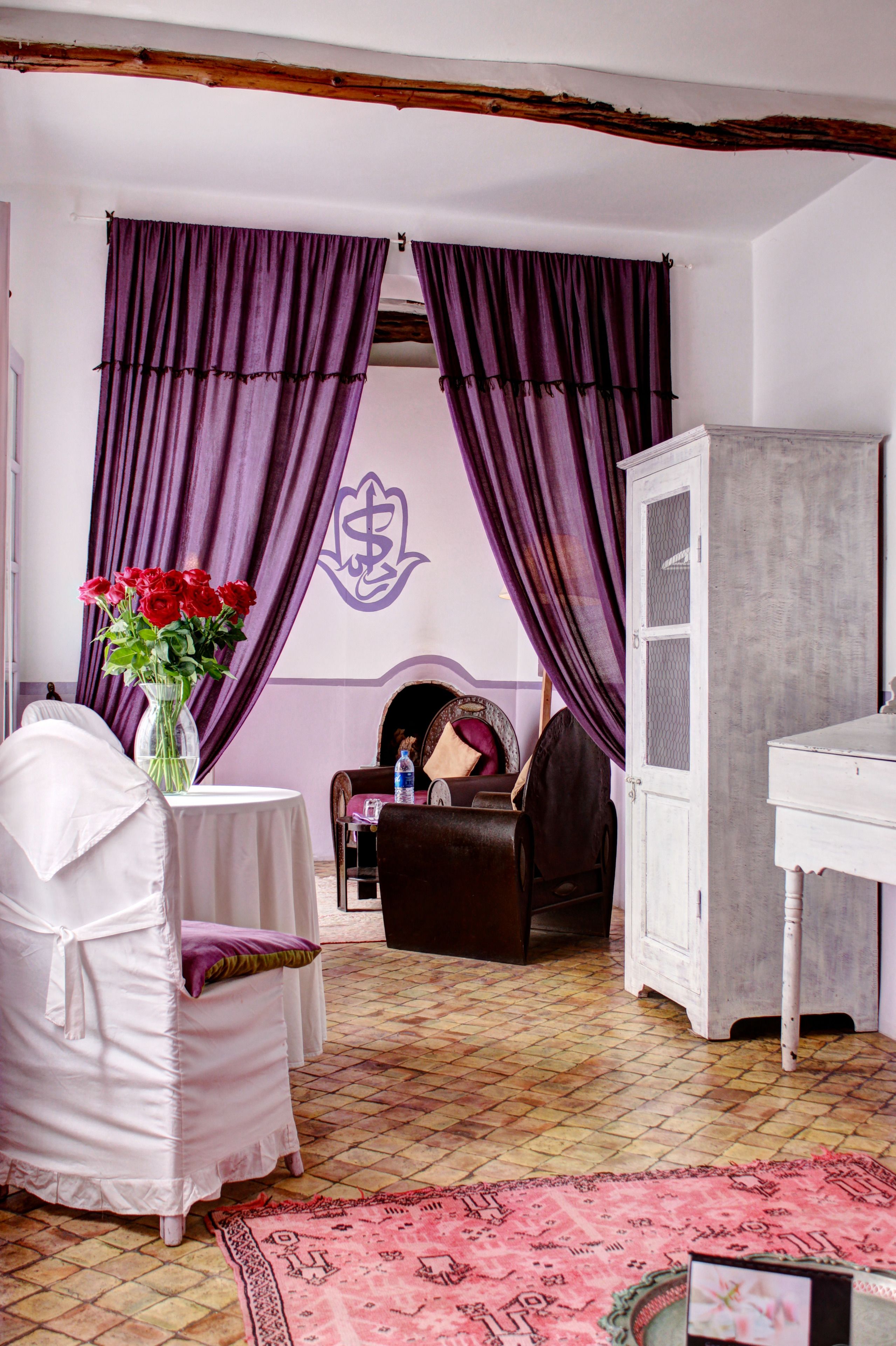 A room with a white chair, a table with a vase of red roses, purple curtains, a fireplace, and a white cabinet.