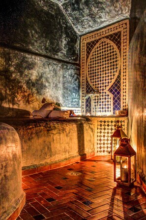 Una habitación tenuemente iluminada con un ventanal decorativo, un farol y un banco con cojines.