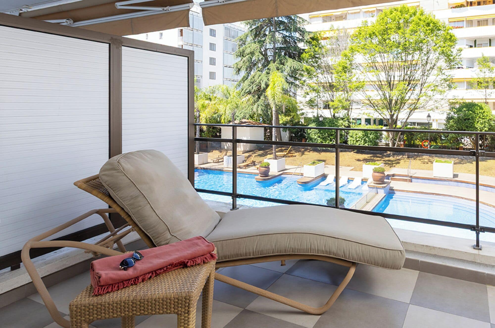 A balcony with a wicker chair, a red cushion, and a pair of sunglasses. A pool with multiple sections and a view of surrounding buildings and trees is visible through the glass.