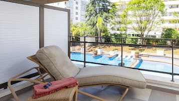 A balcony with a wicker chair, a red cushion, and a pair of sunglasses. A pool with multiple sections and a view of surrounding buildings and trees is visible through the glass.