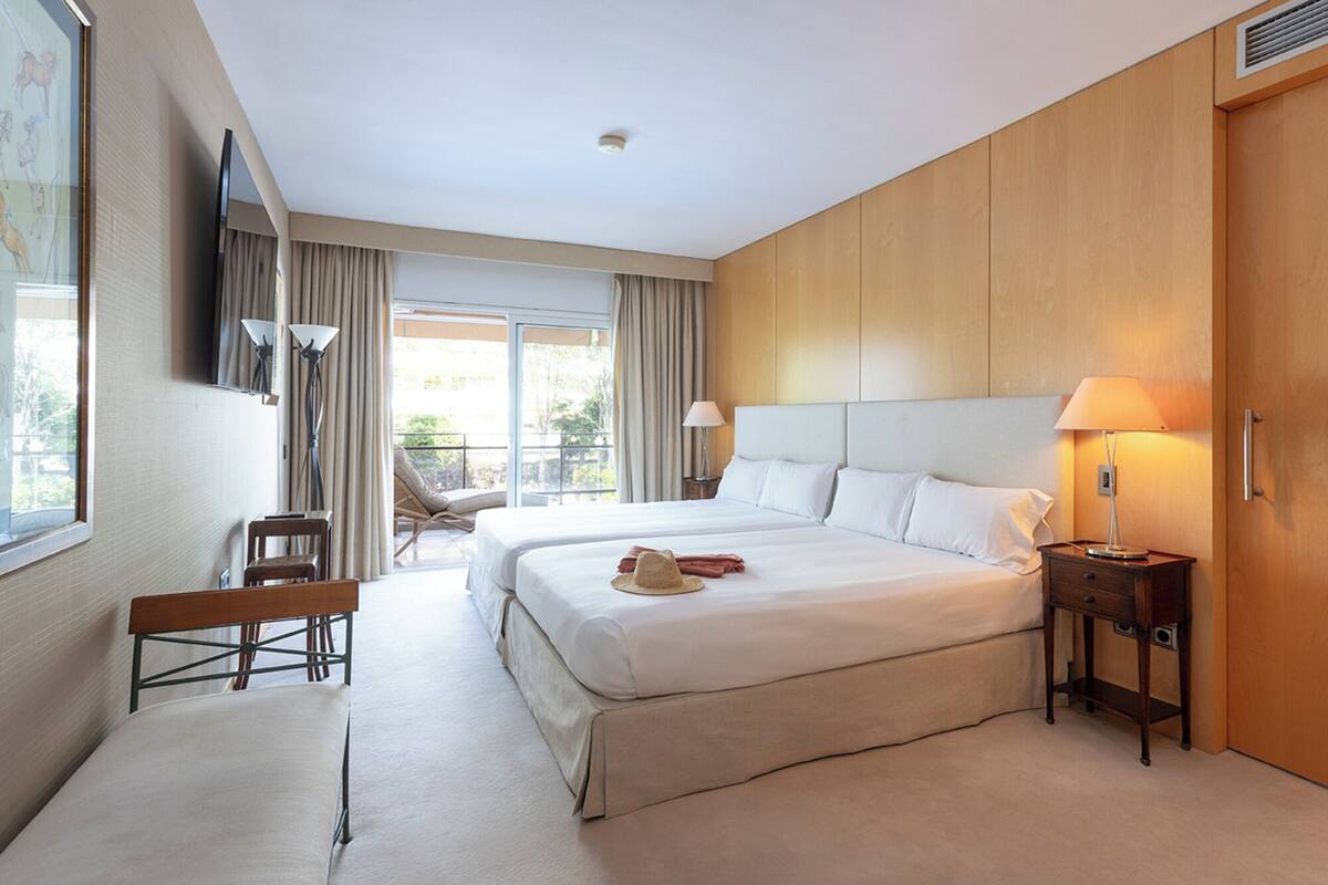 Une chambre d’hôtel avec deux lits, un banc, une chaise, une télévision et une table de chevet avec une lampe.