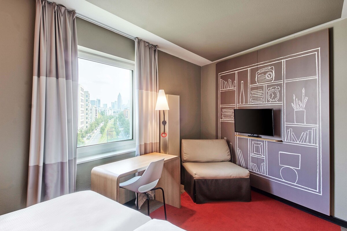 A hotel room with a bed, a desk, a chair, a TV, and a city view.
