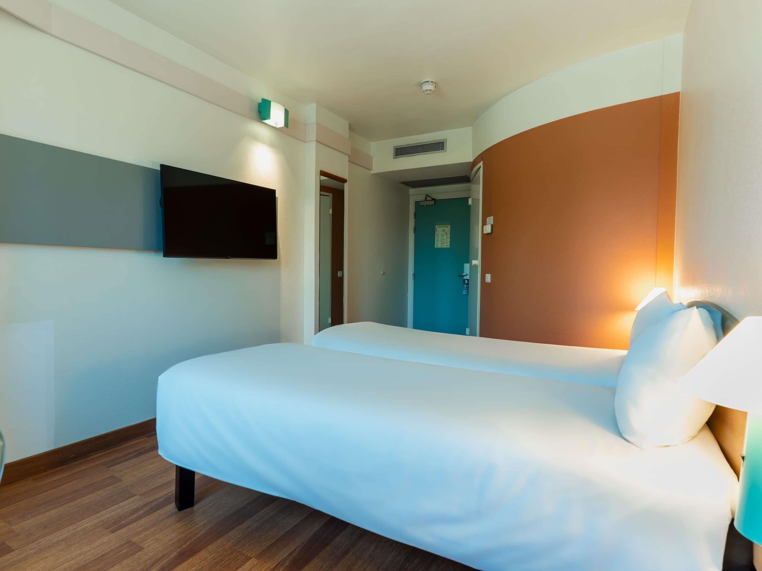Habitación de hotel con una cama, un televisor de pantalla plana sujeto a la pared, una puerta y una lámpara fijada a la pared.