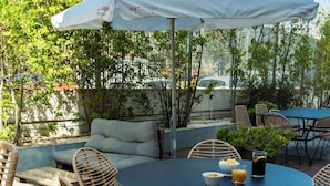 Un patio al aire libre con una mesa redonda, sillas de mimbre, un paraguas blanco y follaje.