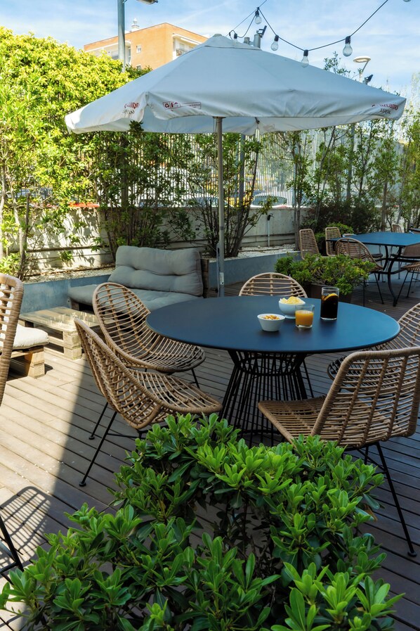 An outdoor patio with a round table, wicker chairs, a white umbrella, and greenery.