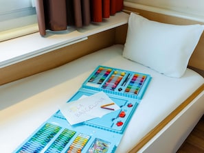 Un lit avec un plateau bleu contenant des crayons de couleur et des crayons à papier, un oreiller blanc et un mot posé sur le lit.