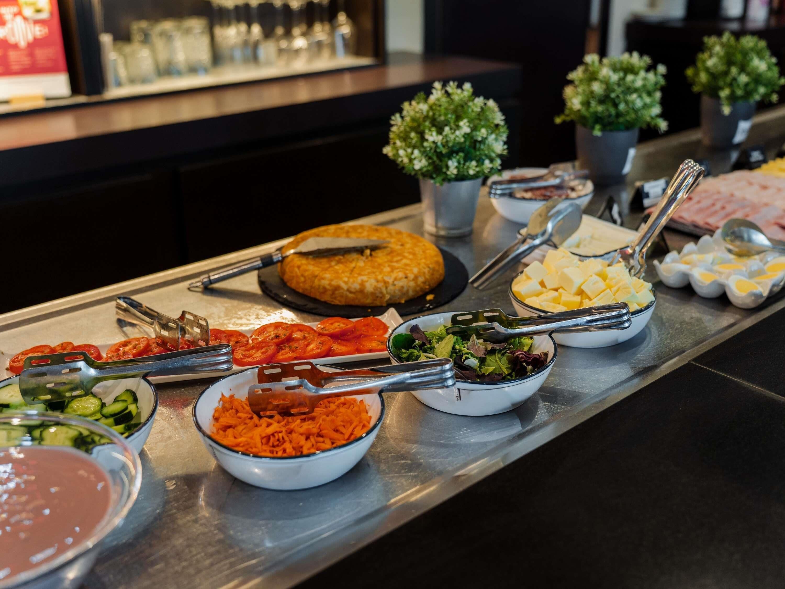 A buffet table with various food items, including sliced tomatoes, shredded carrots, a salad, cheese cubes, and boiled eggs.