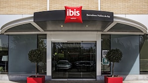 El exterior del hotel ibis Barcelona Molins de Rei, con un diseño moderno, destaca por su llamativo logotipo rojo, una amplia entrada de vidrio y dos árboles en macetas al frente.