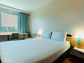 Une chambre d’hôtel avec un grand lit, un bureau, une chaise, deux lampes de chevet et une fenêtre avec des rideaux.