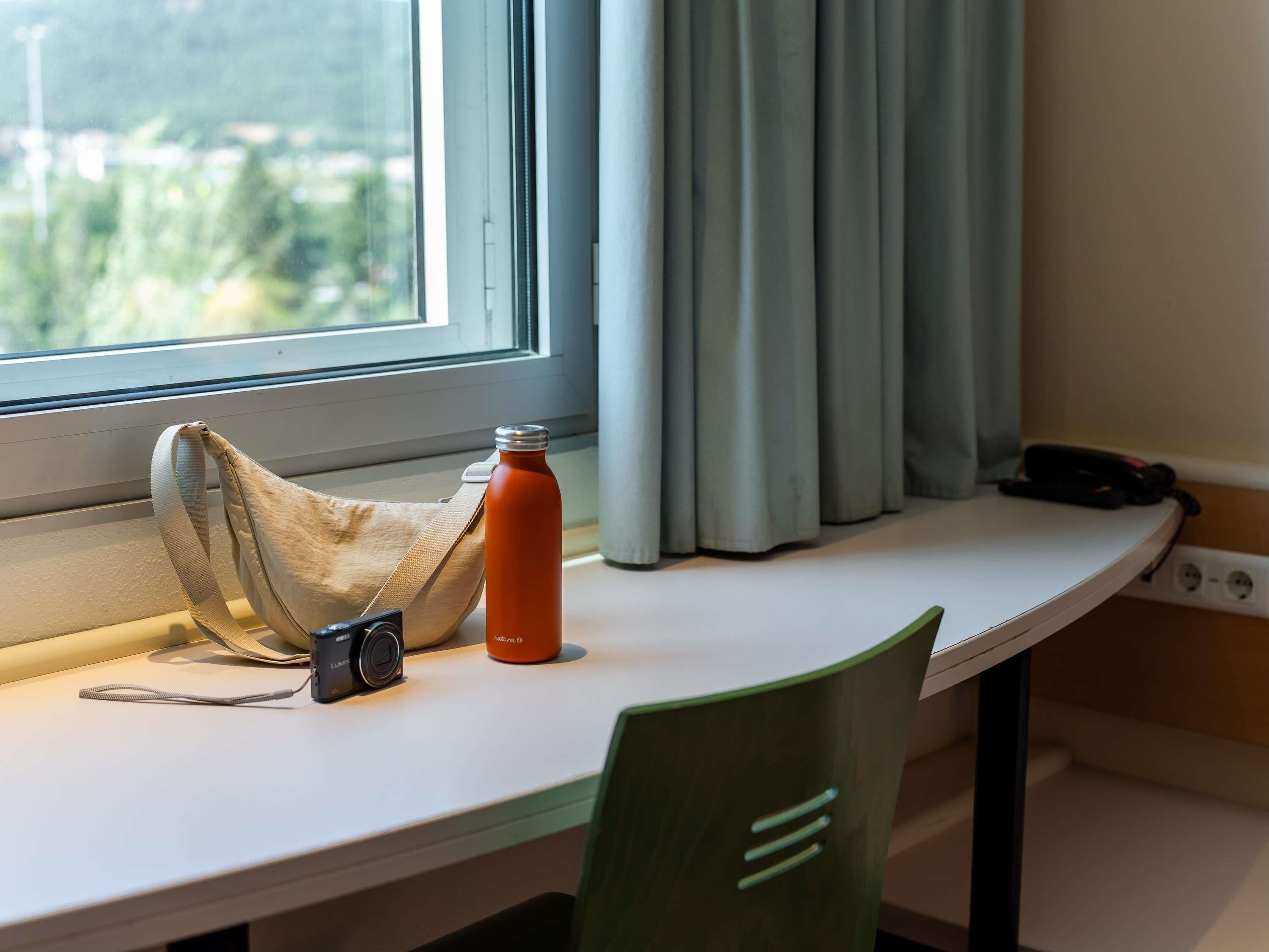 A window with curtains, a table with a camera, a water bottle, and a chair.