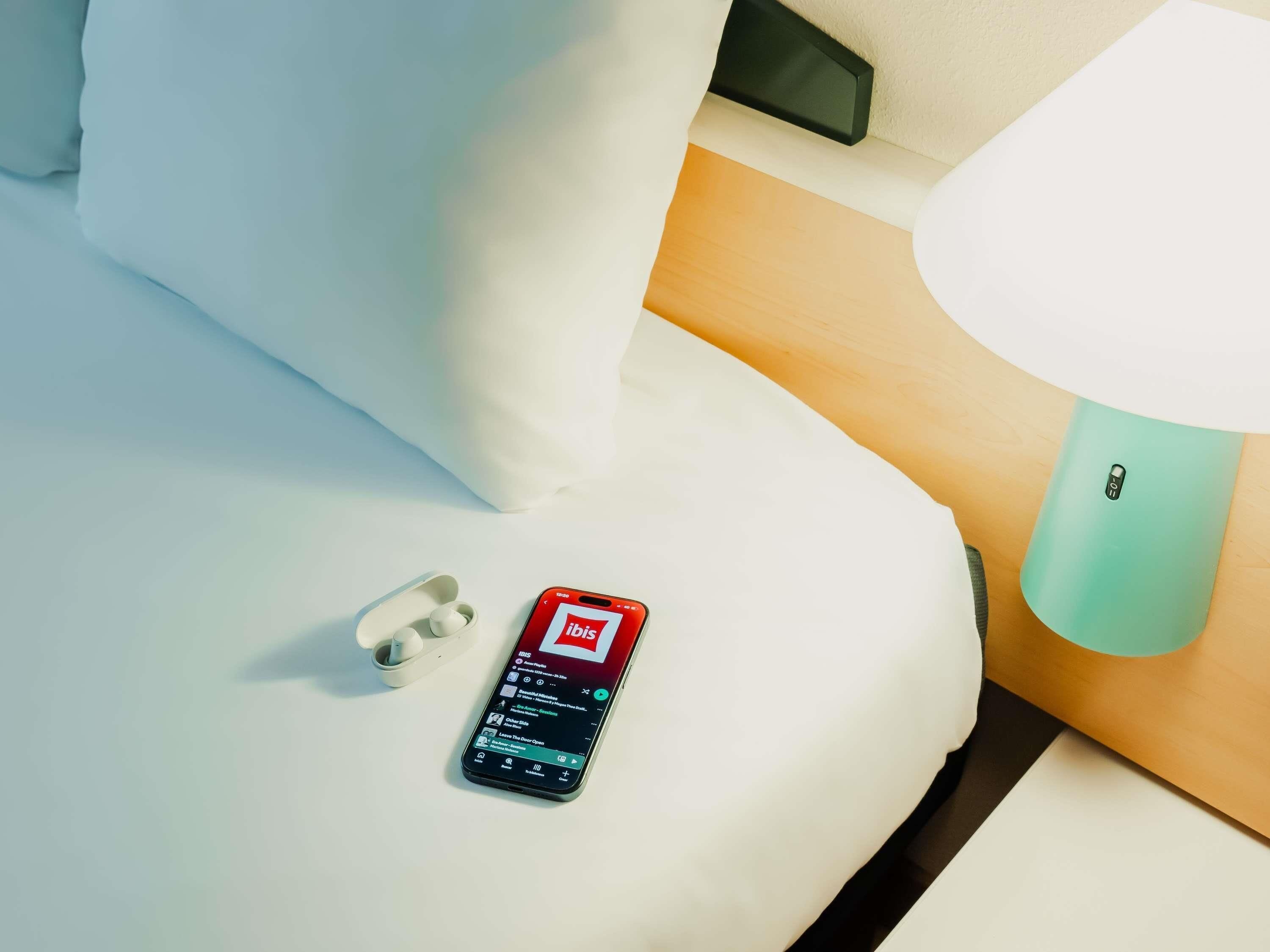 Um smartphone sobre uma cama com um travesseiro branco e uma luminária azul-turquesa.