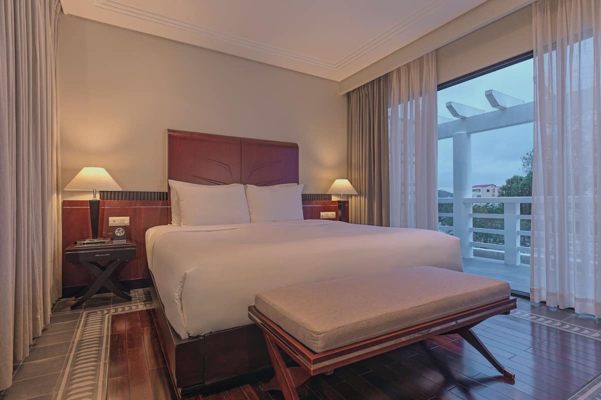 A hotel room with a large bed, two bedside tables with lamps, a wooden floor, and a view of the outside.