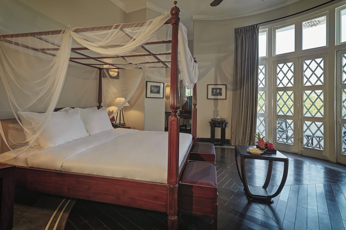 A four-poster bed with a canopy, a bedside table with a lamp, and a view through a window with a lattice design.