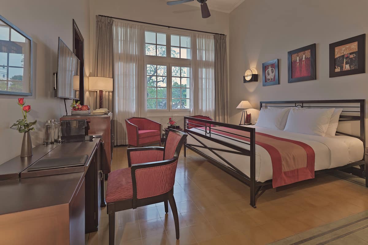 A hotel room with a bed, two chairs, a desk, a ceiling fan, and large windows with curtains.