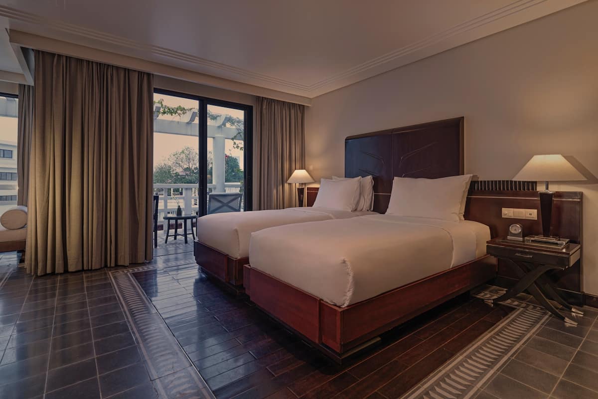 A hotel room with two beds, a large window with curtains, a small table, and a nightstand with a lamp.