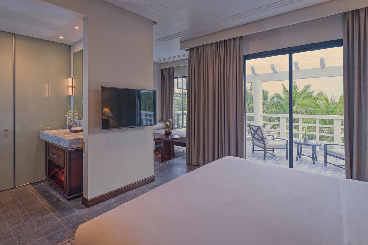 A hotel room with a bed, a TV, a bathroom, and a balcony with a view of palm trees.