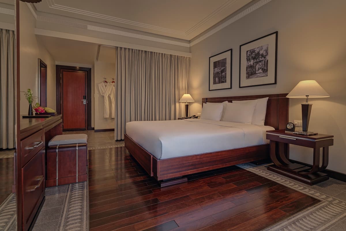 A hotel room with a large bed, bedside lamps, a wooden nightstand, a desk with a chair, a wardrobe, and a bathroom visible in the background.