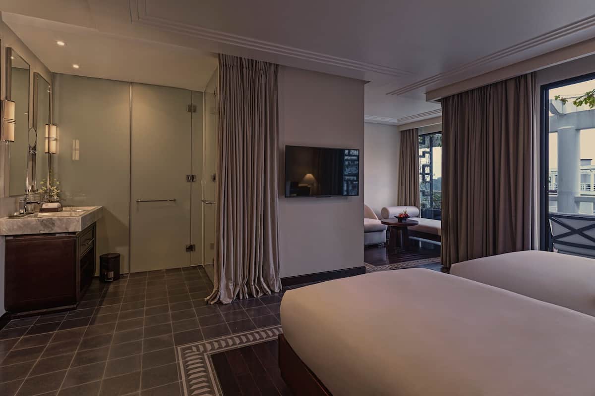 A hotel room with a large bed, a TV mounted on the wall, a bathroom with a mirror and sink, and a balcony with curtains.