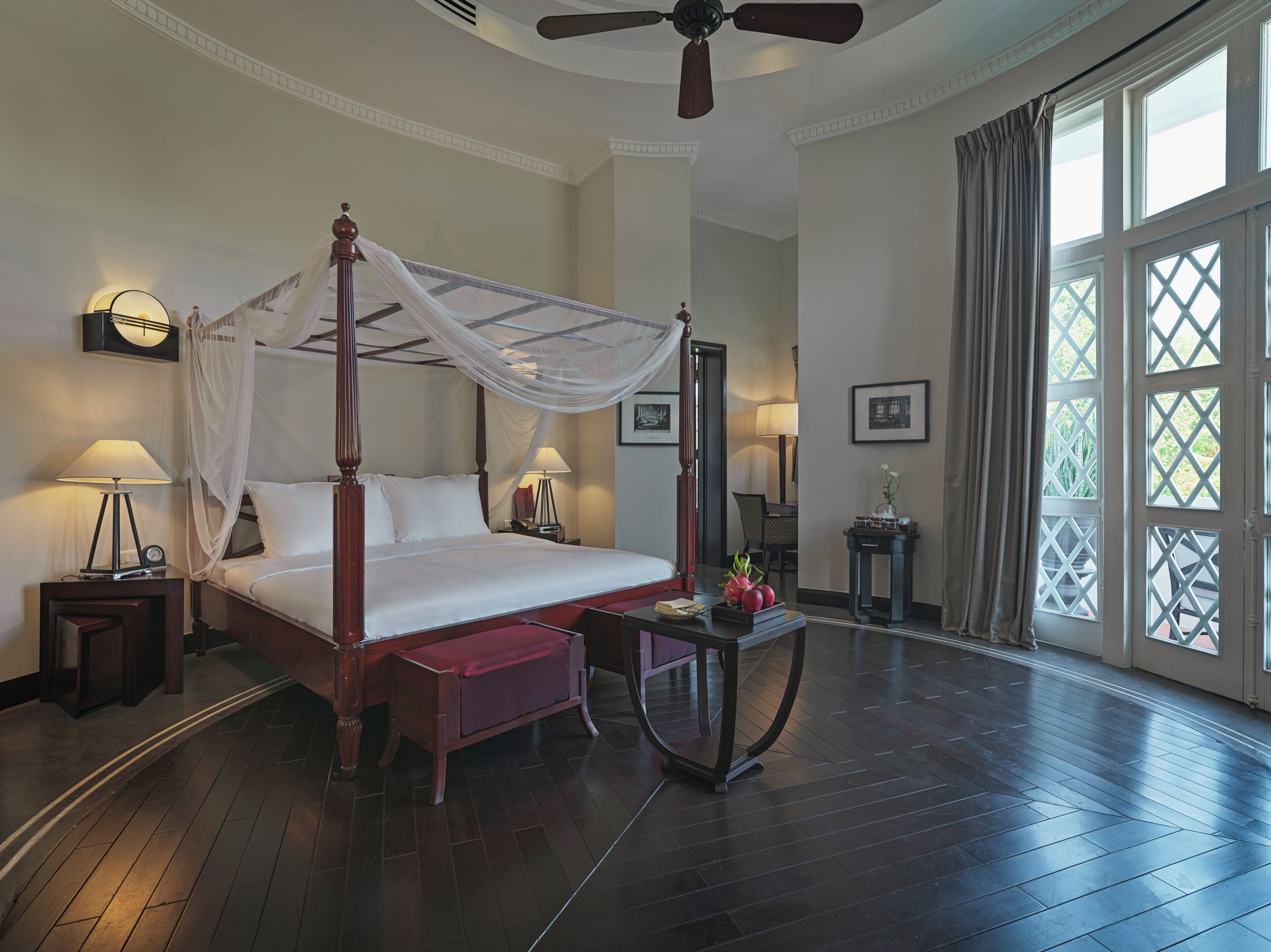 A four-poster bed with a canopy, bedside lamps, a small table with a vase, and a view of the outdoors through large windows.