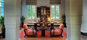 A room with a wooden desk, two chairs, a bookshelf, and two windows with a view of greenery.