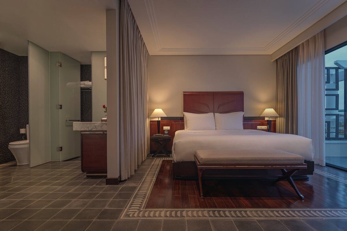 A hotel room with a large bed, a bedside table, a nightstand, a bathroom with a toilet, and a view of the outside.