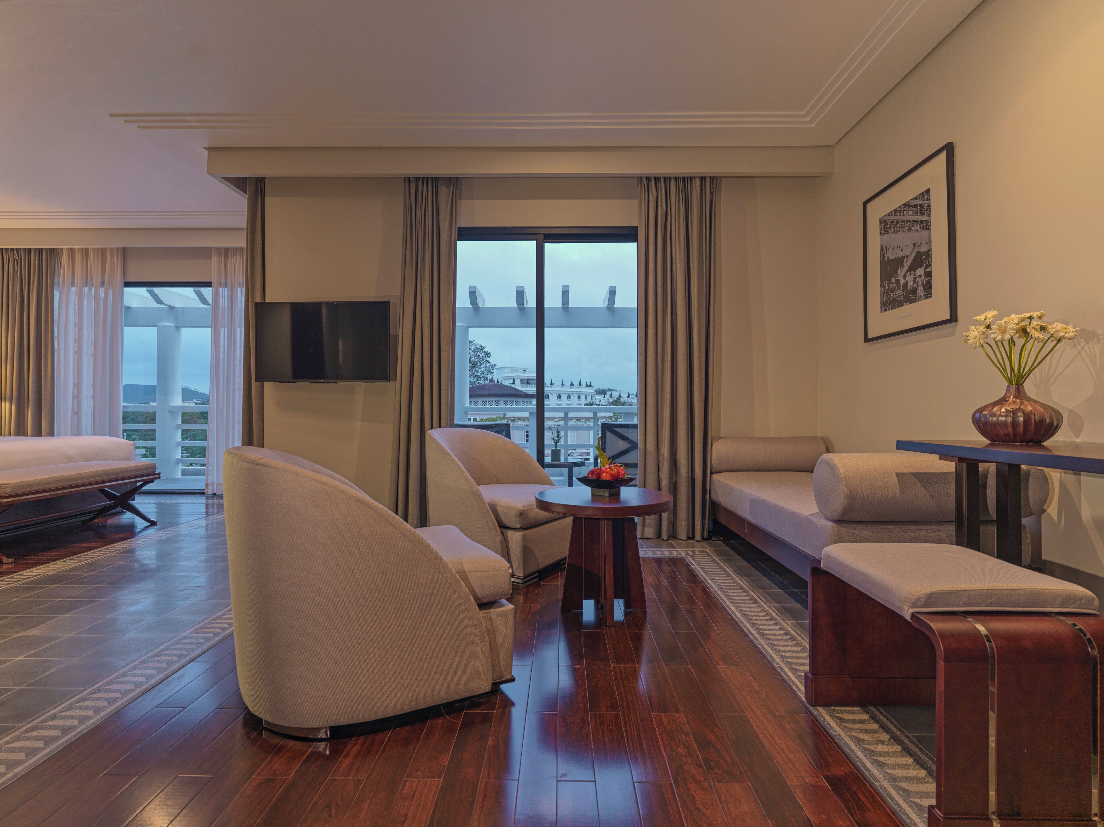 A hotel room with a sofa, armchairs, a small table, and a view of a cityscape through large windows.