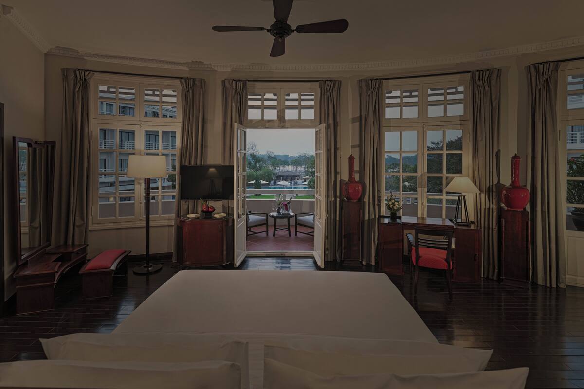A hotel room with a large bed, a TV, a desk with a lamp, a chair, and a view of the outdoors through a large window.