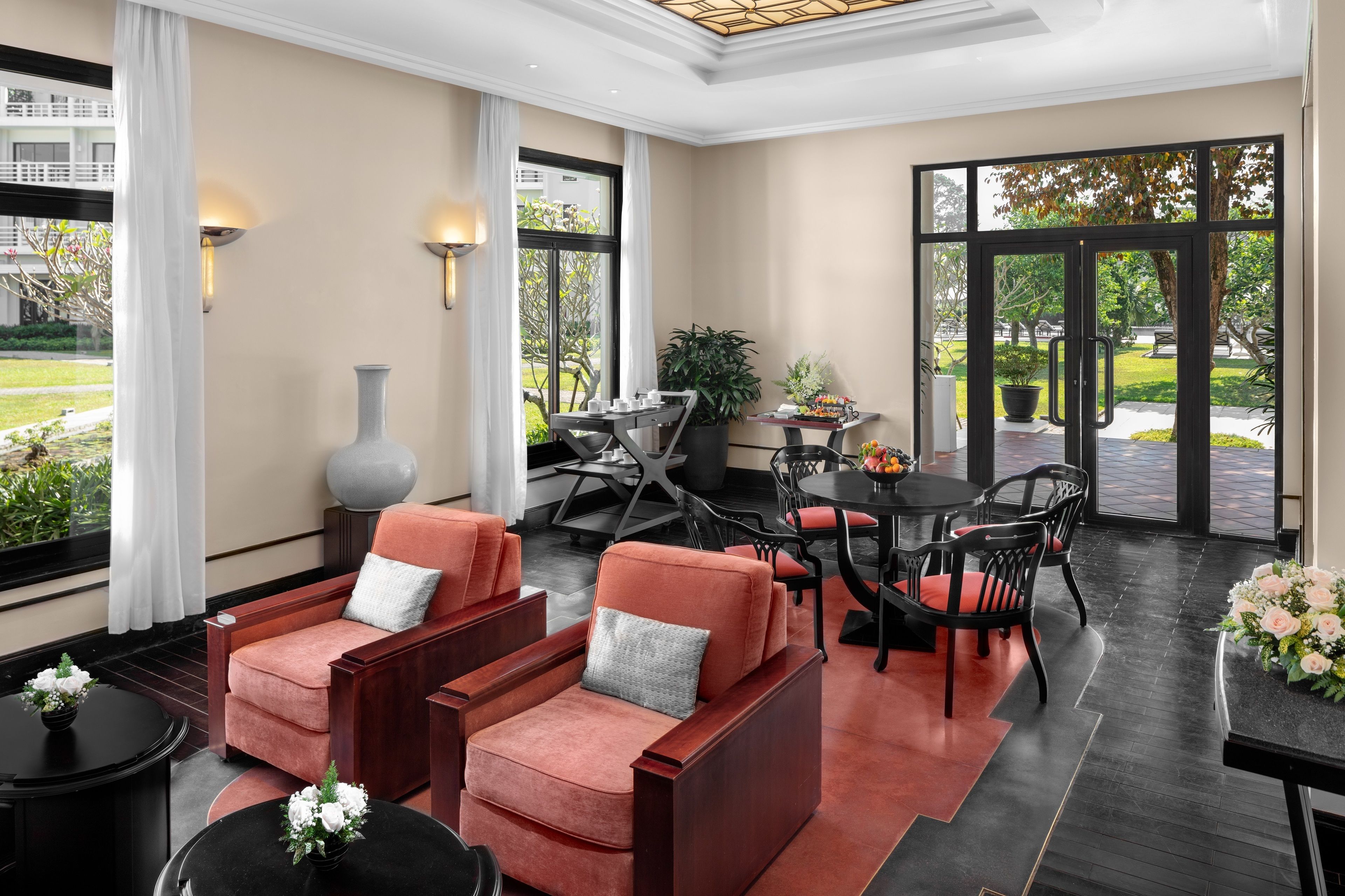 A modern lounge area with two red armchairs, a round table, and a dining table with chairs. Large windows provide a view of the outdoors.