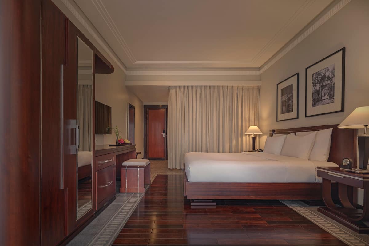 A hotel room with a large bed, a wooden wardrobe, a desk with a chair, a small stool, a nightstand with a lamp, and two framed pictures on the wall.