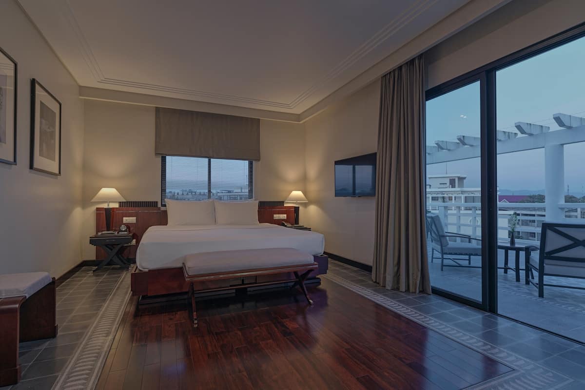 A hotel room with a large bed, two bedside tables, a TV, and a balcony with outdoor seating.
