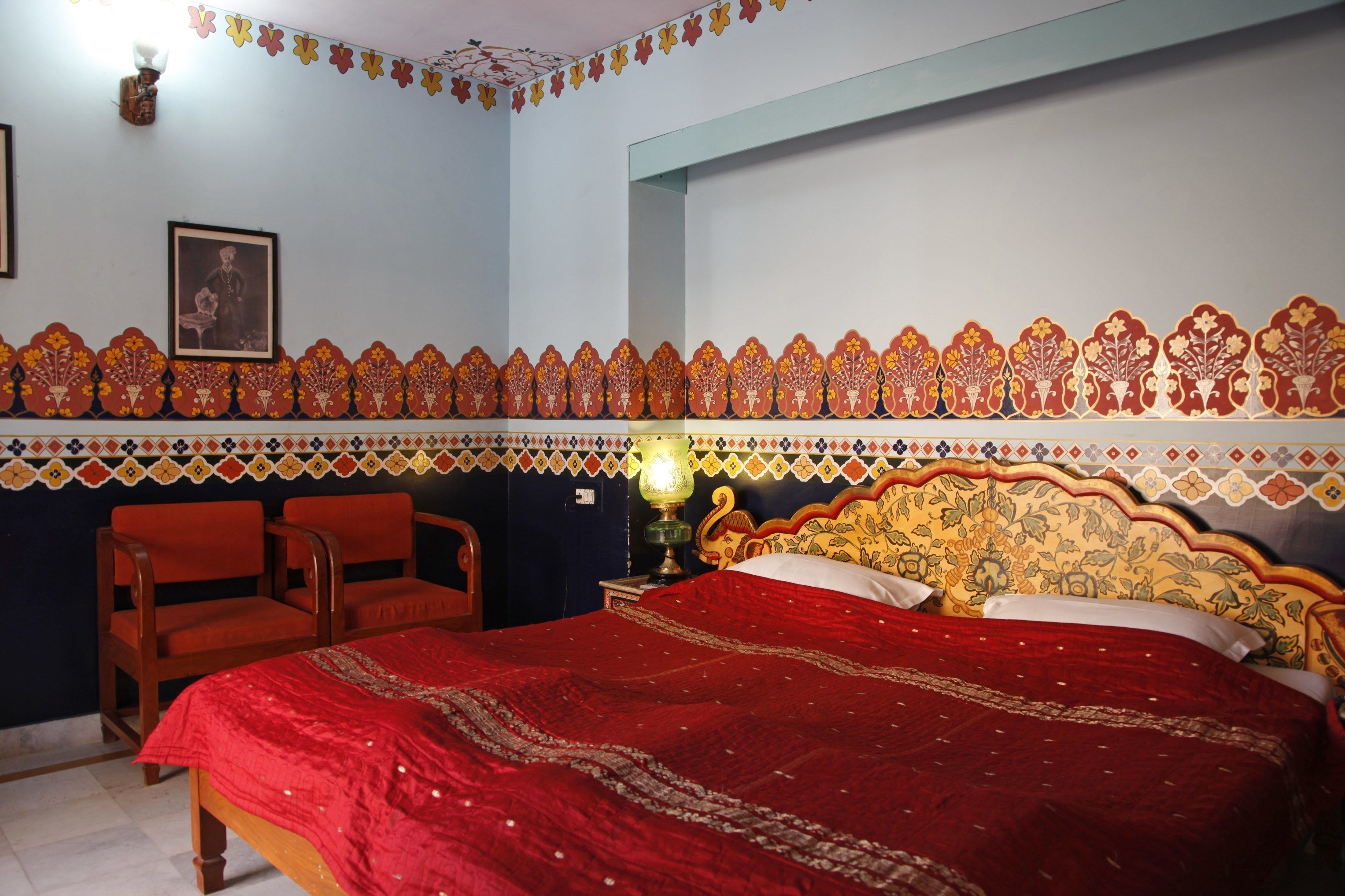 A bedroom with a large bed, two chairs, a nightstand with a lamp, and a wall with decorative patterns.