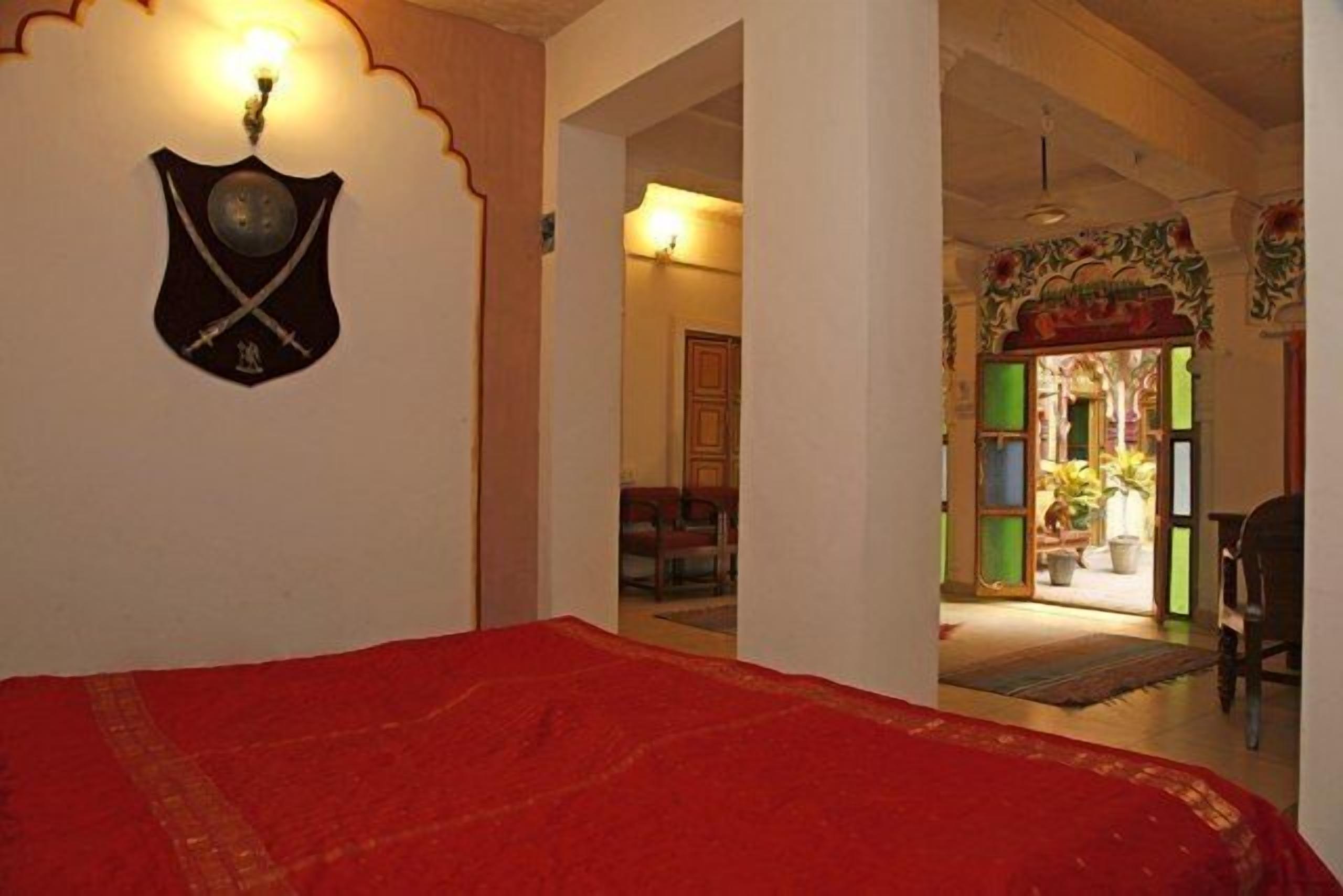 A room with a red bedspread, a shield with crossed swords on the wall, and a doorway leading to a balcony with plants.