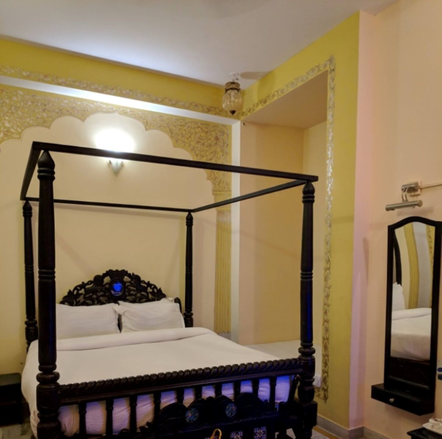 Ein Bett mit Baldachin, mit weißer Bettdecke und schwarzem Gestell, einem an der Wand befestigten Spiegel und einer dekorativen Deckenleuchte.