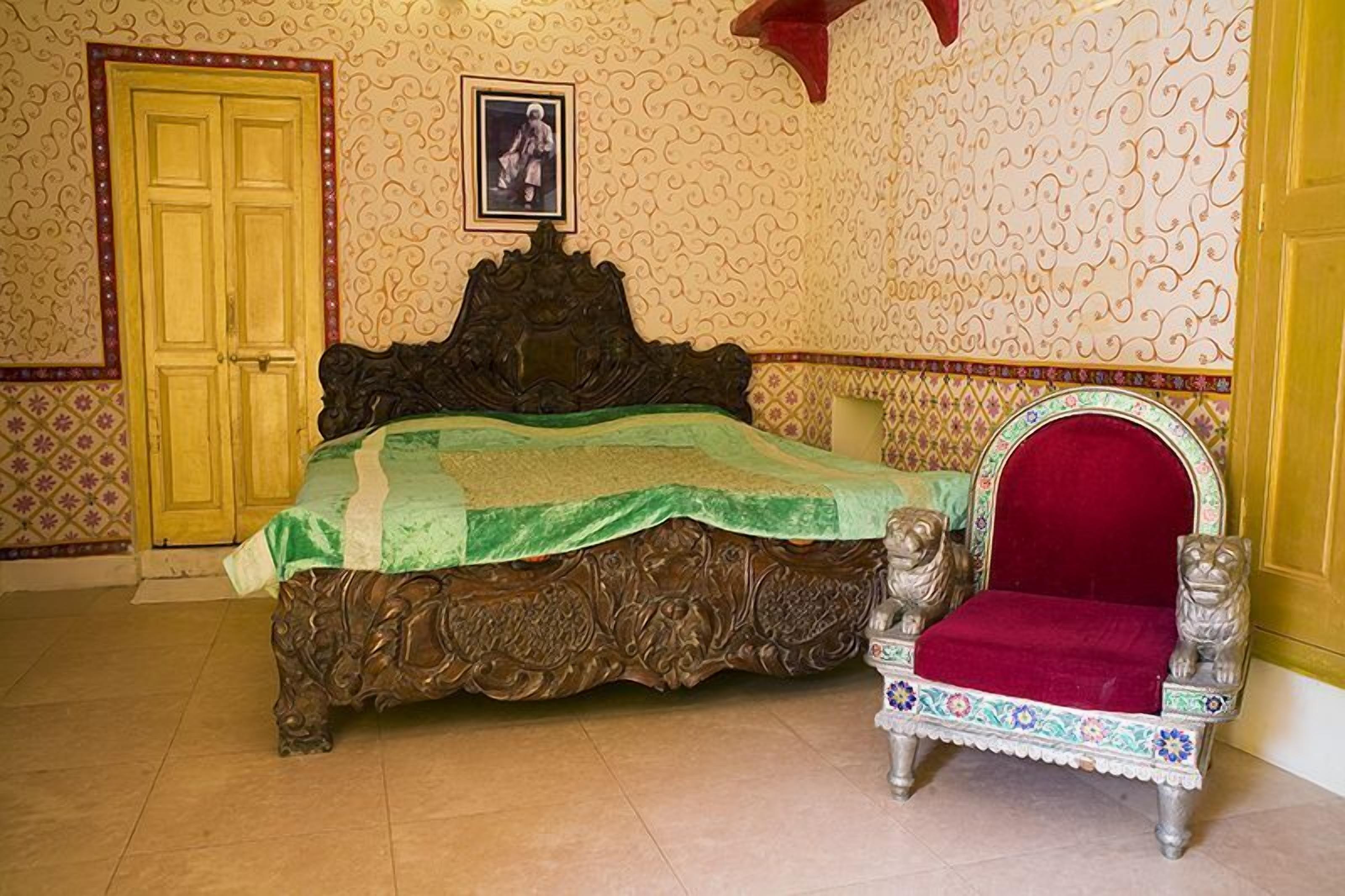 Una habitación con una cama grande y decorativa, una silla ornamental y papel tapiz estampado.
