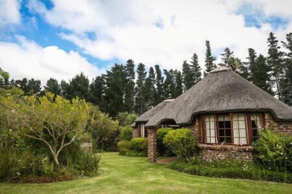 A traditional thatched-roof house with a stone foundation, surrounded by lush greenery and tall trees.