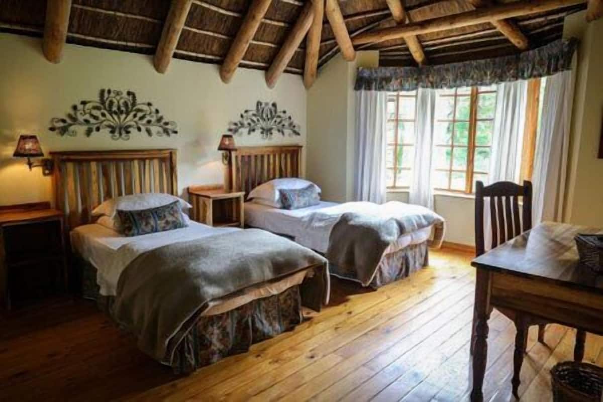 A room with two beds, wooden floors, a table, and a window with curtains.